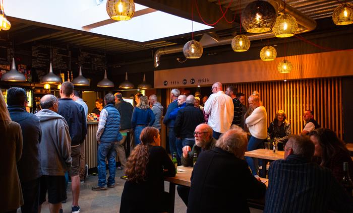 People stand and sit in a warmly light bar area with low hanging lighting and wooden vertical slats towards the rear of the image
